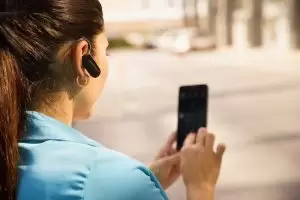 Dışarıda, mavi gömlekli, atkuyruğu saçlı bir kadın, sağ kulağında Siemens Signia Çok Yönlü Telefonda İşitme Cihazı Kullanımı kablosuz kulaklığı takılıyken akıllı telefonunu kullanıyor. Arka plan bulanık.
