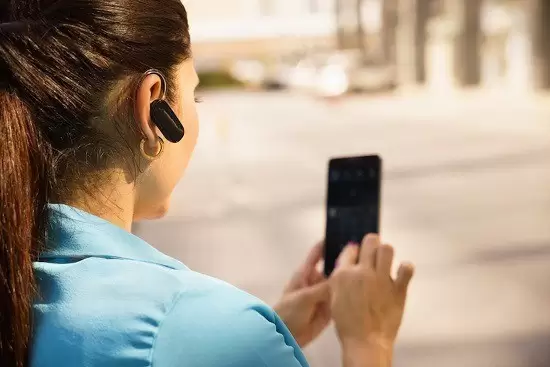Dışarıda, mavi gömlekli, atkuyruğu saçlı bir kadın, sağ kulağında Siemens Signia Çok Yönlü Telefonda İşitme Cihazı Kullanımı kablosuz kulaklığı takılıyken akıllı telefonunu kullanıyor. Arka plan bulanık.
